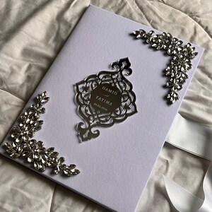 May include: A lilac wedding invitation with silver detailing. The invitation has a decorative silver centerpiece with the names "Hamid" and "Fatima" and the date "01.02.2026". Silver floral accents are on the corners, and a white ribbon is visible.