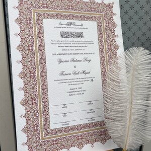 Op de afbeelding: Een huwelijkscertificaat met sierlijke bordeauxrode en gouden randdetails. Het document bevat Arabische schrift en Engelse tekst, die een huwelijk bevestigt. Een witte veren pen ligt in de buurt, wat duidt op een formele gelegenheid.