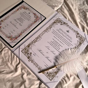 K&ouml;nnte beinhalten: Ein offener Hochzeitsurkundentr&auml;ger mit zwei Dokumenten, mit floralen R&auml;ndern und kunstvoller Kalligraphie. Eine wei&szlig;e Federkiel und ein goldener Stift liegen auf dem Dokument. Die Dokumente befinden sich auf wei&szlig;em Hintergrund.