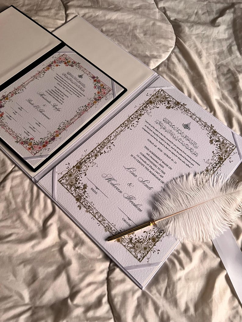 K&ouml;nnte beinhalten: Ein offener Hochzeitsurkundentr&auml;ger mit zwei Dokumenten, mit floralen R&auml;ndern und kunstvoller Kalligraphie. Eine wei&szlig;e Federkiel und ein goldener Stift liegen auf dem Dokument. Die Dokumente befinden sich auf wei&szlig;em Hintergrund.