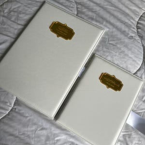 Op de afbeelding: Twee witte ledergebonden boeken met goudkleurige decoratieve labels. De boeken zijn rechthoekig en hebben een gestikte rand. De labels hebben Arabische tekst. E&eacute;n boek is groter dan het andere.