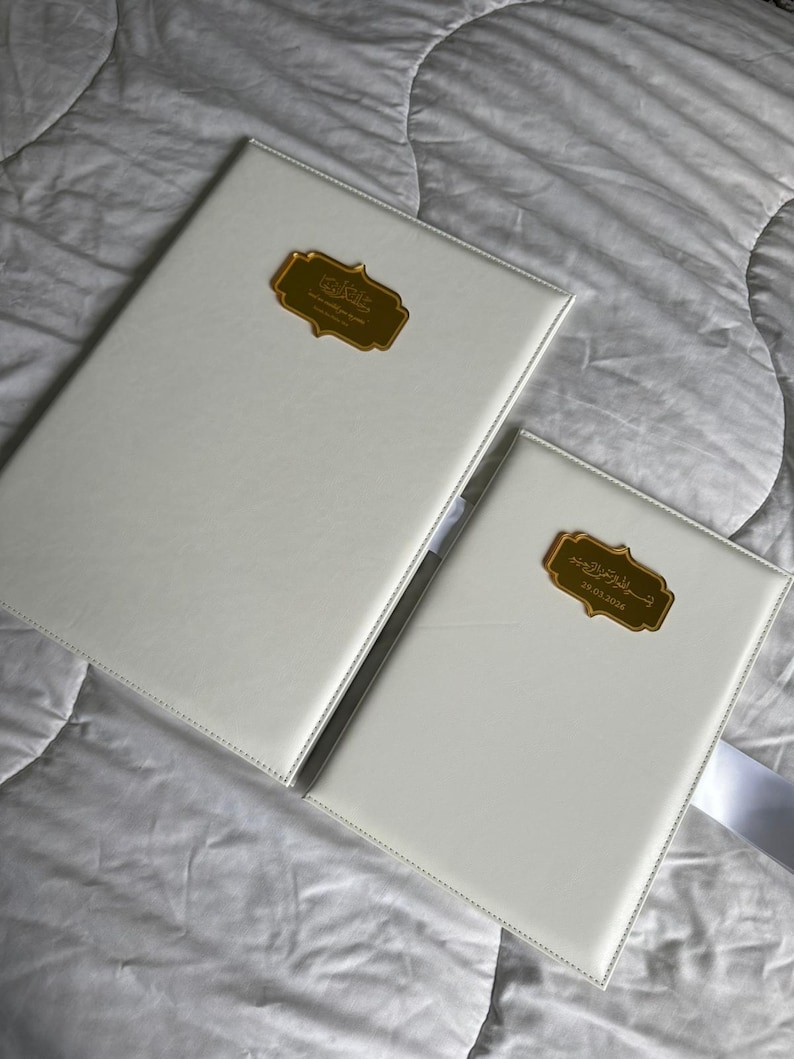 Op de afbeelding: Twee witte ledergebonden boeken met goudkleurige decoratieve labels. De boeken zijn rechthoekig en hebben een gestikte rand. De labels hebben Arabische tekst. E&eacute;n boek is groter dan het andere.