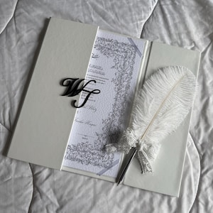 May include: A white wedding invitation set with a decorative feather pen. The invitation is in a white folder with a black monogram. The pen has a white feather and a lace bow. The invitation has text and floral border.