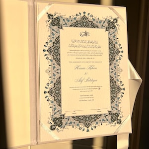 May include: An open, cream-colored wedding certificate with ornate blue and gray floral border. The certificate includes text in Arabic and English, with the names Hania Khan and Atif Siddique. A white ribbon is attached.