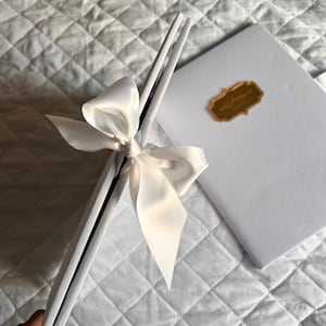 K&ouml;nnte beinhalten: Ein wei&szlig;es Buch und eine Reihe wei&szlig;er, rechteckiger Gegenst&auml;nde sind mit einer gro&szlig;en, cremefarbenen Satinschleife zusammengebunden. Auf dem Buch befindet sich ein goldener Aufkleber mit Text. Die Artikel liegen auf einer wei&szlig;en, gesteppten Oberfl&auml;che.