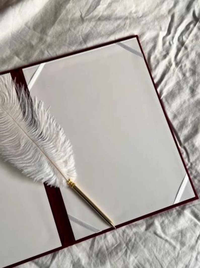 Op de afbeelding: Een open boek met bordeauxrode randen en witte pagina's, geaccentueerd met witte hoekhouders van lint. Een witte ganzenveerpen met een goudkleurige metalen punt ligt op de open pagina's, wat wijst op een thema van schrijven of documentatie.