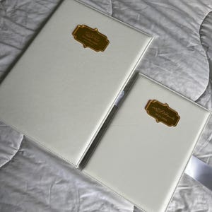 K&ouml;nnte beinhalten: Zwei wei&szlig; ledergebundene B&uuml;cher mit goldfarbenen rechteckigen Etiketten. Die Etiketten haben arabische Schrift. Die B&uuml;cher liegen auf einer wei&szlig;en, strukturierten Oberfl&auml;che. Ein Buch ist gr&ouml;&szlig;er als das andere.
