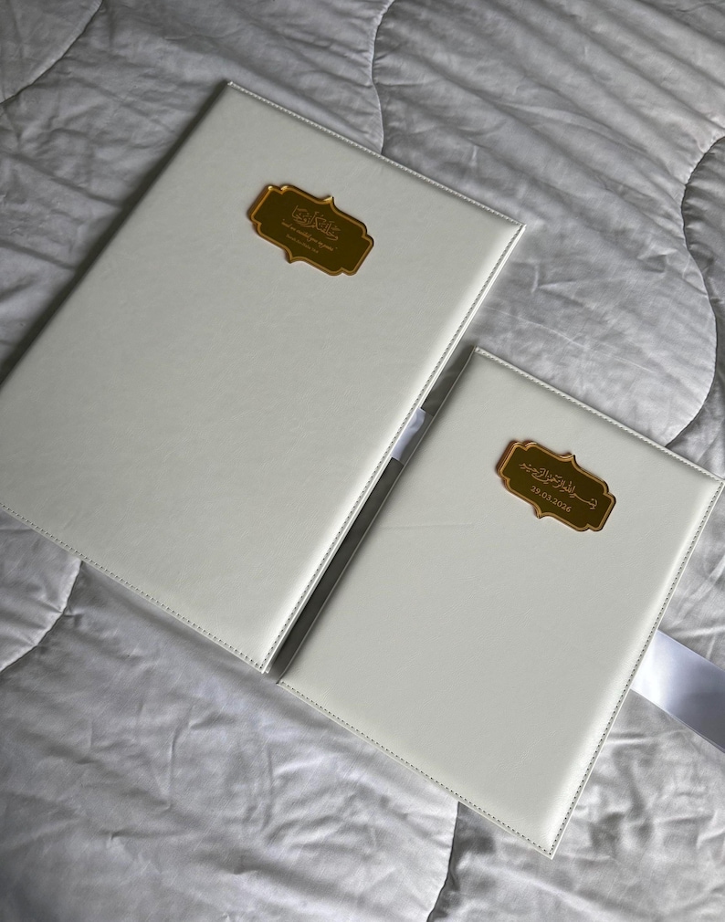 K&ouml;nnte beinhalten: Zwei wei&szlig; ledergebundene B&uuml;cher mit goldfarbenen rechteckigen Etiketten. Die Etiketten haben arabische Schrift. Die B&uuml;cher liegen auf einer wei&szlig;en, strukturierten Oberfl&auml;che. Ein Buch ist gr&ouml;&szlig;er als das andere.