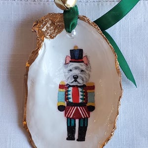 May include: A unique Christmas ornament made from a white oyster shell with a gold-painted edge. The shell features a colorful illustration of a dog dressed as a nutcracker, with a green ribbon and gold bell for hanging.