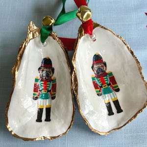 May include: Two decorative oyster shells with gold-painted edges, each featuring a painted nutcracker soldier with a pug dog face. The shells are suspended by green and red ribbons, each adorned with a small gold jingle bell at the top attachment.
