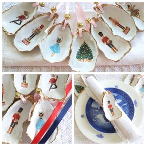 May include: A collection of white, shell-shaped ornaments with gold edges, featuring festive illustrations of a Christmas tree, a ballerina, and a toy soldier. Each ornament is adorned with a pink ribbon and a small gold bell, perfect for holiday decor.