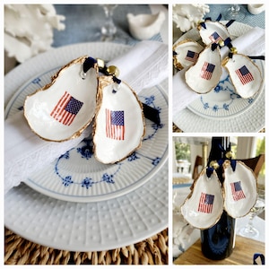 May include: White oyster shell ornaments with a gold rim and an American flag design. Each shell is adorned with a navy blue ribbon and a small gold bell. The ornaments are displayed on white plates with blue floral patterns.