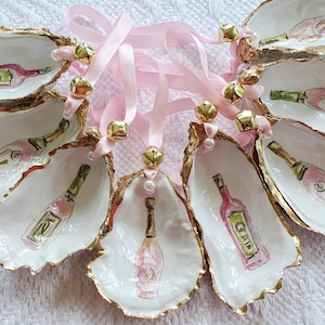 May include: Eight oyster shell ornaments with gold trim and pink ribbon. Each shell features a hand-painted design of a bottle of champagne or wine.