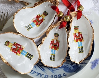 Puede incluir: Cinco conchas de ostra blancas con ribete dorado, cada una decorada con un diseño de cascanueces diferente. Las conchas están dispuestas en un plato blanco con un patrón azul y blanco.