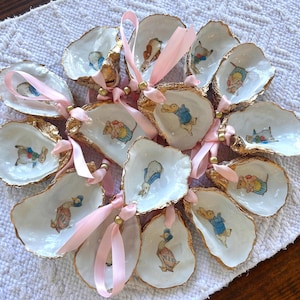 May include: A collection of 12 oyster shells painted with gold accents and various illustrations of Peter Rabbit characters. Each shell has a pink ribbon and gold bead attached.