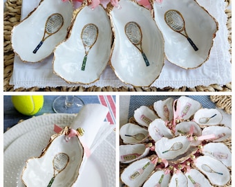 May include: A collection of oyster shell ornaments featuring hand-painted tennis rackets on the interior, with gold-painted edges and pink ribbon loops with small gold bells attached to the top of each shell for hanging or decorative use.