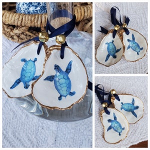 May include: Two white oyster shell ornaments with gold trim and blue painted sea turtles. The ornaments are hanging from a blue ribbon with gold bells.