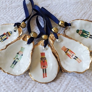 May include: Five oyster shell ornaments with gold trim and nutcracker illustrations. Each ornament has a blue ribbon with a gold bell attached.
