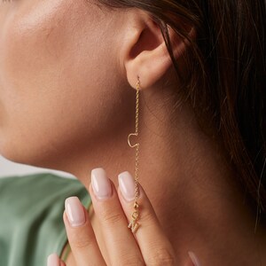 May include: A gold-colored, long chain earring with the letter "C" at the top and the letter "M" at the bottom, adorned with small, sparkling stones. The earring is dangling from an ear, showcasing a delicate and personalized jewelry piece.