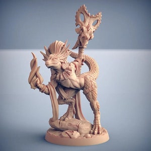 May include: A miniature figurine of a lizard-like creature with a staff and a flame in its hand. The creature is wearing a robe and has a detailed, scaly body. The figurine is painted in a light brown color.