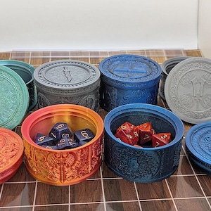 Pu&ograve; includere: Una collezione di contenitori rotondi dal design ornato in vari colori, tra cui verde acqua, arancione, blu e grigio. Alcuni contenitori sono aperti, rivelando dadi. I coperchi presentano dettagliati motivi a rilievo.