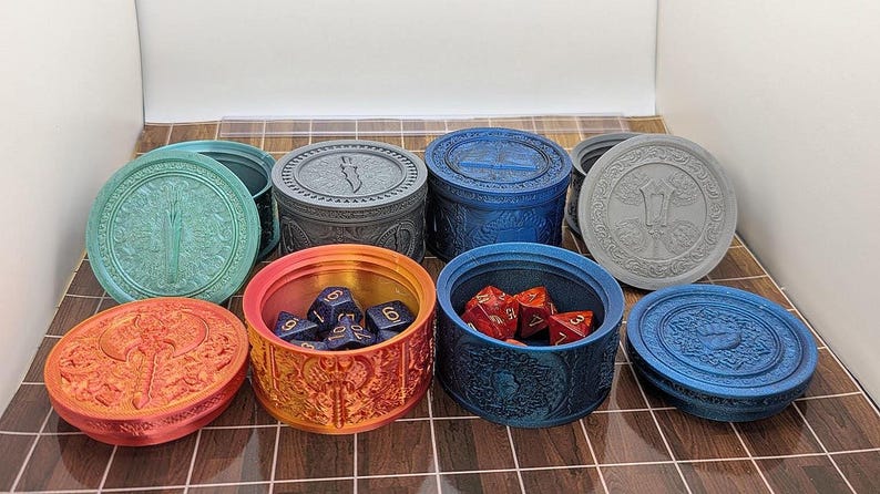 Pu&ograve; includere: Una collezione di contenitori rotondi dal design ornato in vari colori, tra cui verde acqua, arancione, blu e grigio. Alcuni contenitori sono aperti, rivelando dadi. I coperchi presentano dettagliati motivi a rilievo.