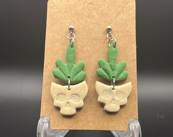 Puede incluir: Un par de pendientes colgantes con un cráneo blanco y detalles de plantas verdes. Los pendientes se muestran en una tarjeta marrón.