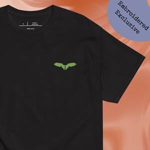 Può includere: T-shirt nera con un piccolo disegno di ali verdi ricamate sul petto. La maglietta è realizzata in 100% cotone. Le parole "Embroidered Exclusive" sono stampate su un cerchio blu in alto a destra.