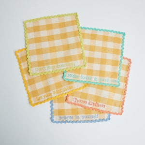 May include: Five square, yellow and white gingham fabric patches with scalloped edges in various colors. Each patch has an embroidered inspirational phrase: "Don't stop dreaming," "Make today a good one," "Let your light shine," "Choose kindness," and "Believe in yourself."