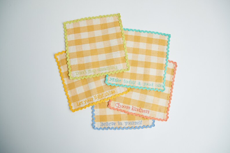 May include: Five square, yellow and white gingham fabric patches with scalloped edges in various colors. Each patch has an embroidered inspirational phrase: "Don't stop dreaming," "Make today a good one," "Let your light shine," "Choose kindness," and "Believe in yourself."
