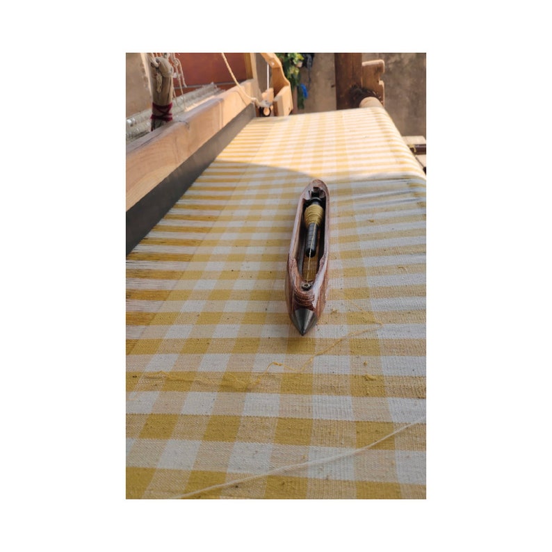 May include: A wooden weaving shuttle with a metal tip rests on a yellow and white checkered surface. The shuttle is brown with a silver tip and contains a spool of yellow thread. The background includes a wooden loom.