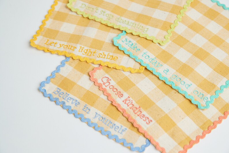 May include: Close-up of several yellow and white checkered fabric squares with embroidered inspirational quotes. Each square has a different colored scalloped edge and embroidered text, including "Don't stop dreaming" and "Choose Kindness."