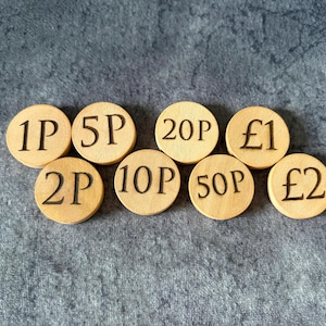 May include: A set of eight circular light-coloured wooden tokens, each engraved with a different British currency denomination: 1P, 2P, 5P, 10P, 20P, 50P, £1, and £2. The tokens are arranged in a cluster, displaying clear black text for each value.