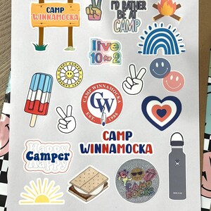 May include: A collection of colorful stickers featuring camp-themed designs, including a peace sign, a campfire, a rainbow, a smiley face, a popsicle, a heart, a sun, a s'more, a water bottle, and a sign that reads "Camp Winnamocka".