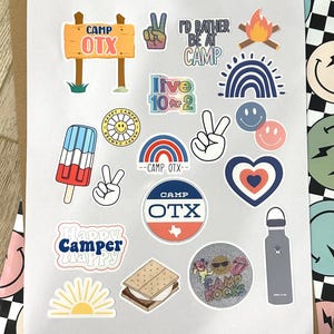 May include: A sheet of stickers with a camp theme. The stickers feature a variety of designs, including a sign that says "Camp OTX", a peace sign, a campfire, a rainbow, a popsicle, a heart, a s'more, a sun, and a water bottle. The stickers are perfect for adding a touch of summer fun to your belongings.