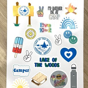May include: A sheet of stickers with a camping theme. The stickers feature images of a campfire, a rainbow, a peace sign, a heart, a s'more, a water bottle, a sun, a popsicle, and a logo for "Lake of the Woods".