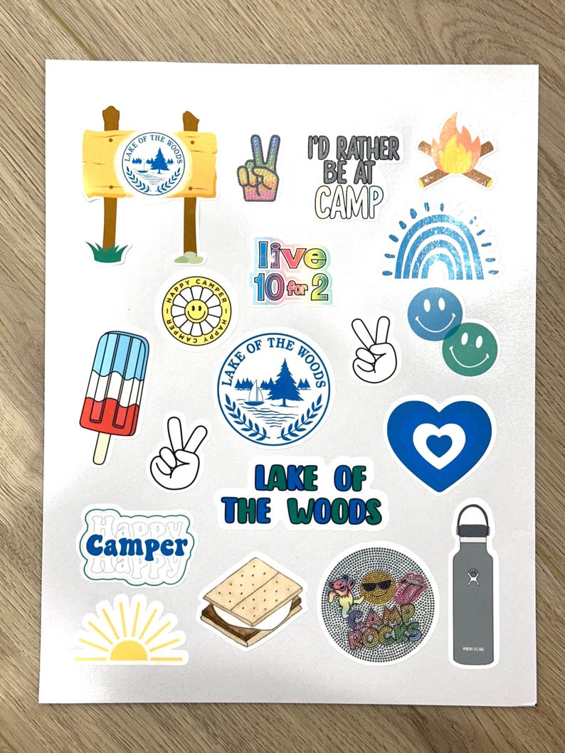 May include: A sheet of stickers with a camping theme. The stickers feature images of a campfire, a rainbow, a peace sign, a heart, a s'more, a water bottle, a sun, a popsicle, and a logo for "Lake of the Woods".