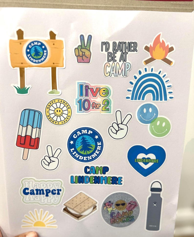 May include: A collection of colorful stickers featuring camp-themed designs, including a peace sign, a campfire, a rainbow, a smiley face, a popsicle, a heart, a sign that says "Camp Lindenmere", and a s'more.