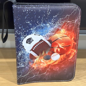 Puede incluir: Una bolsa negra con cremallera con temática deportiva. La bolsa presenta un balón de fútbol, un balón de baloncesto, un balón de béisbol y un disco de hockey en un diseño de fuego con salpicaduras de agua.