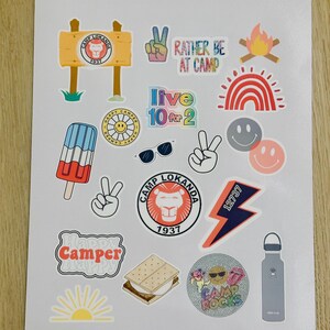 May include: A sheet of colorful stickers with a camp theme. The stickers feature a variety of designs, including a lion, a campfire, a rainbow, a smiley face, a peace sign, a popsicle, sunglasses, a lightning bolt, a s'more, a sun, and a water bottle. The text on the stickers includes "Camp Lokanda", "Rather Be At Camp", "Live 10 for 2", "Happy Camper", and "Camp Rocks".