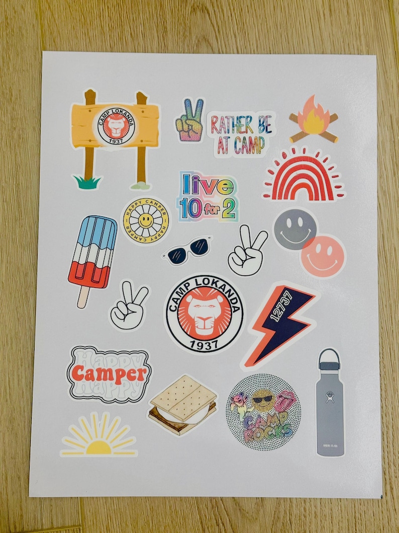 May include: A sheet of colorful stickers with a camp theme. The stickers feature a variety of designs, including a lion, a campfire, a rainbow, a smiley face, a peace sign, a popsicle, sunglasses, a lightning bolt, a s'more, a sun, and a water bottle. The text on the stickers includes "Camp Lokanda", "Rather Be At Camp", "Live 10 for 2", "Happy Camper", and "Camp Rocks".