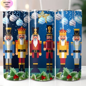 May include: Three stainless steel tumblers with a blue background and a Christmas nutcracker theme. The tumblers feature four different nutcrackers in red, yellow, blue, and green. The background is decorated with snowflakes and ornaments.