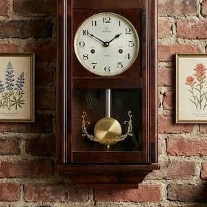 Könnte beinhalten: Eine Wanduhr aus dunklem Holz mit weißem Zifferblatt und schwarzen Zeigern. Die Uhr hat ein Pendel und ist an einer roten Backsteinmauer befestigt. Die Uhr ist im klassischen Vintage-Stil gehalten.