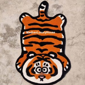Peut inclure: Un tapis en peluche orange et noir en forme de tigre avec un visage blanc et des yeux noirs. Le tapis est à l'envers, la queue pointant vers le haut. Le texte "MadhavLuxuryHouse" est imprimé sur le tapis.