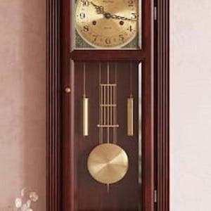 May include: A wall-mounted wooden clock with a dark brown finish, featuring a gold-toned circular dial with black numerals and hands. The lower section contains a glass panel displaying a gold-coloured pendulum and two vertical weights.