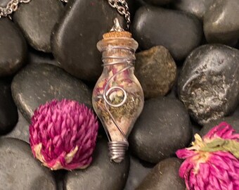 May include: A silver chain necklace with a small glass bottle pendant filled with dried flowers and greenery. The bottle is wrapped with silver wire and has a cork stopper.