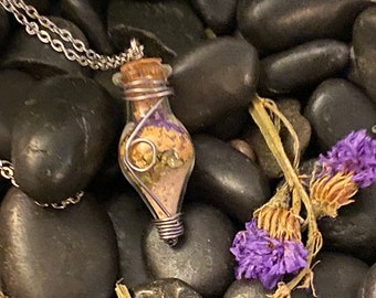 Könnte beinhalten: Eine silberne Kettenkette mit einem kleinen Glasflaschenanhänger. Die Flasche ist mit getrockneten Blumen und Sand gefüllt und mit Silberdraht umwickelt. Die Kette ist auf einem Bett aus schwarzen Steinen fotografiert.