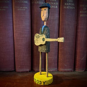 May include: A carved wooden figure of a man playing a guitar, with the words "This Machine Kills Fascists" on the guitar. The figure is wearing a hat and a suit. The base is yellow with the name "Woody". The background features a row of old books.
