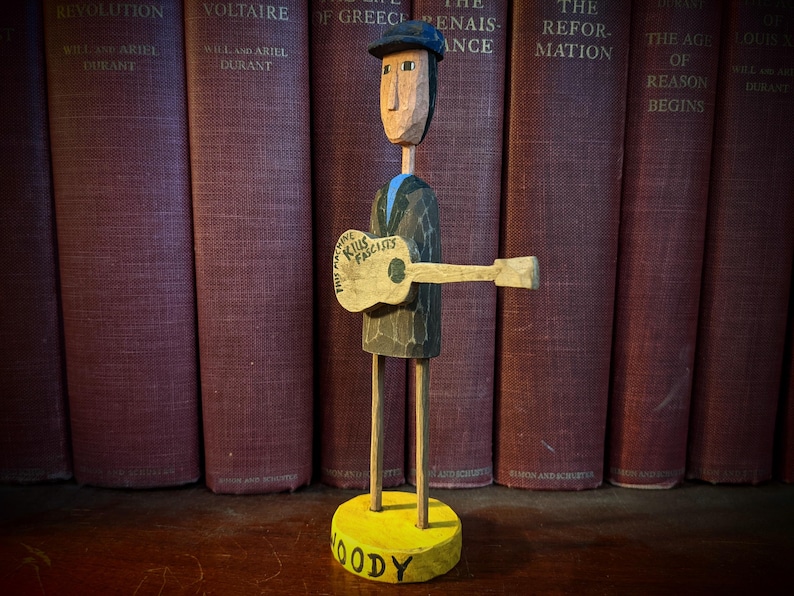 May include: A carved wooden figure of a man playing a guitar, with the words "This Machine Kills Fascists" on the guitar. The figure is wearing a hat and a suit. The base is yellow with the name "Woody". The background features a row of old books.