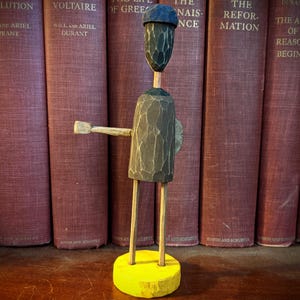 May include: A carved wooden figure with a dark gray body, a blue hat, and a yellow base. The figure has outstretched arms and is positioned in front of a row of vintage books with maroon covers.